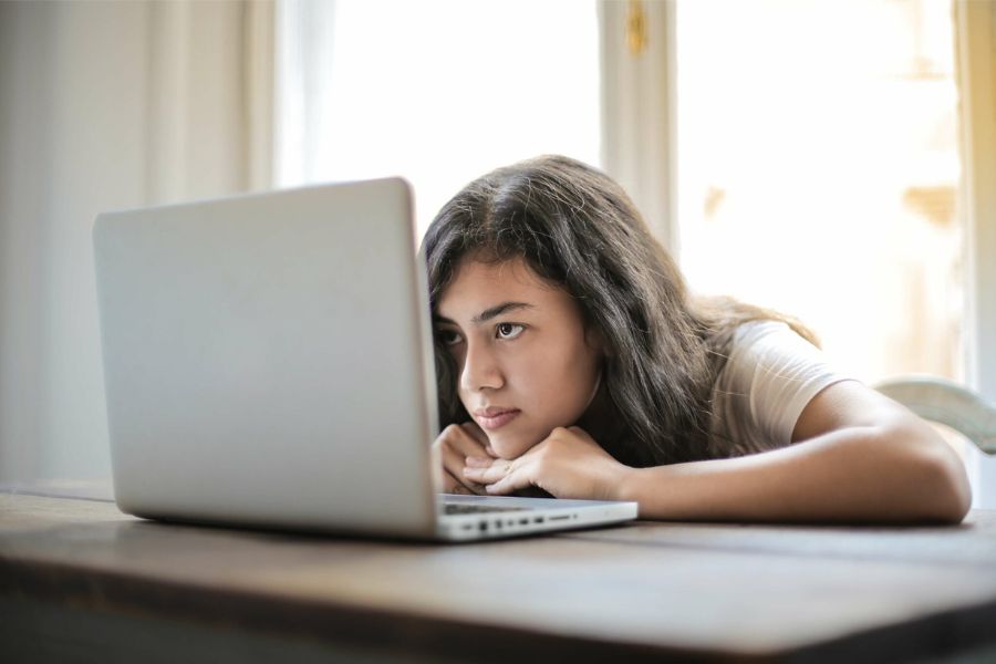Young woman procrastinating by setting her chin on her desk in front of a laptop.