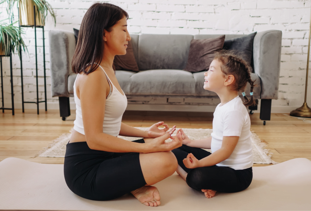 A mother is teaching her daugther the power of meditation for children as they sit on the floor together and relax.