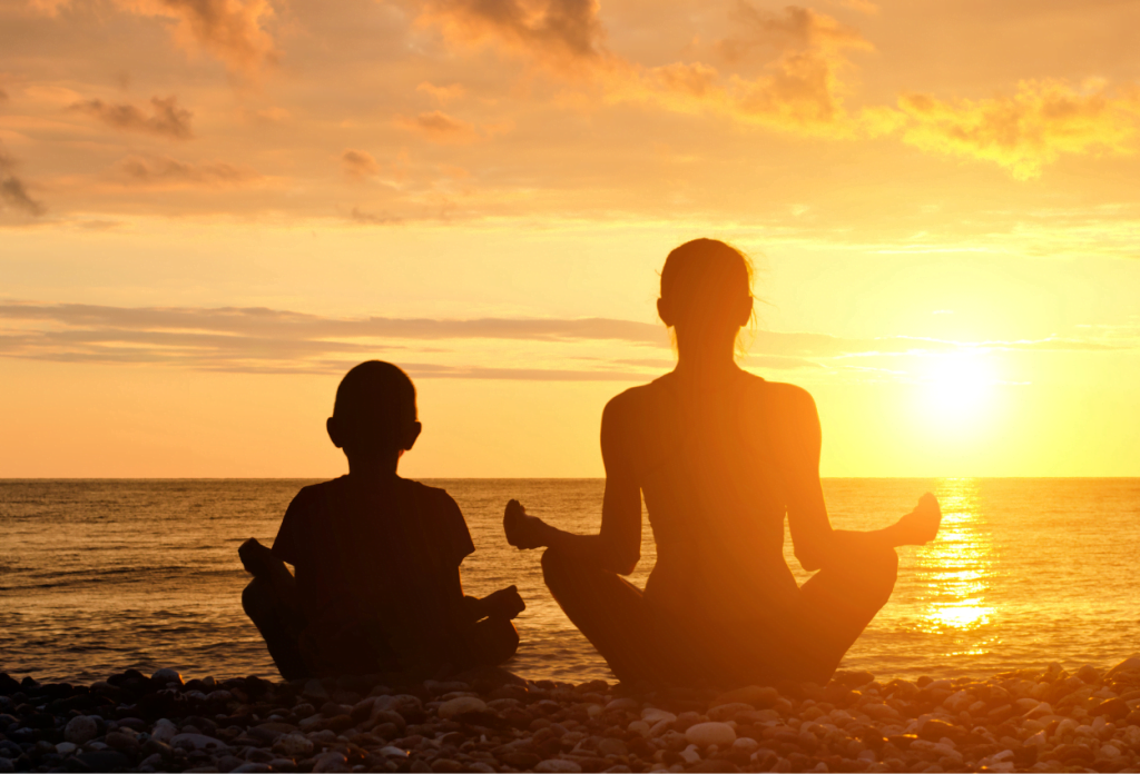 A parent and child are practicing meditation for children while sitting on the beach during sunset.