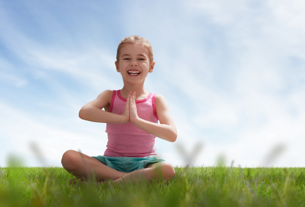 A little girl has her hands folded over her heart and is sitting in the grass practicing meditation for children.
